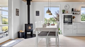 A modern kitchen with white cabinetry, a black wood-burning stove, a dining table with chairs, and a view of greenery outside.