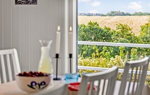 Un'area pranzo con un tavolo apparecchiato per due, una ciotola di frutta e una candela. Una finestra mostra vista su un vigneto e campi.