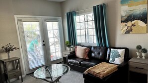 A living room with a black leather sofa, a glass coffee table, and a sliding glass door leading to a balcony. - Studio Steps To The Sand - Spacious, Clean, Cozy (Manhattan Beach)