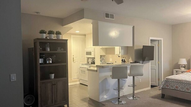 A modern kitchen with white appliances, a dining area with two bar stools, a large bed with a patterned bedspread, and a ceiling fan. - Studio Steps To The Sand - Spacious, Clean, Cozy (Manhattan Beach)