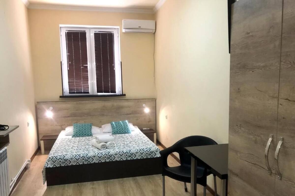 A small, compact room with a bed, bedside table, and a chair. There is a window with blinds, an air conditioning unit, and a wardrobe.