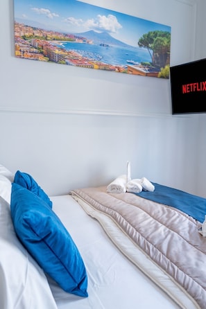 Une chambre d’hôtel avec un lit, un oreiller bleu, une couette beige et blanche, une couverture bleue et un téléviseur fixé au mur qui affiche le logo de Netflix.