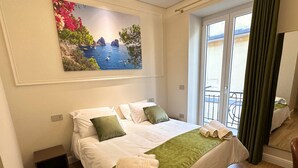 A hotel room with a bed, a painting of a coastal scene, a balcony with a view, and a door. - A CASA TUA  (Naples)