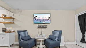 A living room with two blue armchairs, a small white table, a flat-screen TV mounted on the wall, and a white cabinet with shelves. - Oak Shores Studio 4 | Just a Short Walk to the Beach! (Biloxi)