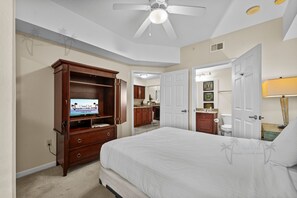 Une chambre à coucher comprenant un lit, une télévision, une armoire, un ventilateur de plafond et une salle de bain visible à travers une porte ouverte.