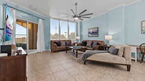 A spacious living room with a ceiling fan, a large window with curtains, a sofa set, a coffee table, and a desk with a chair. - Sienna on the Coast 107 | Beautiful Unit on the Ground Floor! (Gulfport)