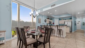A spacious kitchen with a central island, a dining area with a glass table and chairs, and a large window providing a view of greenery. - Sienna on the Coast 107 | Beautiful Unit on the Ground Floor! (Gulfport)