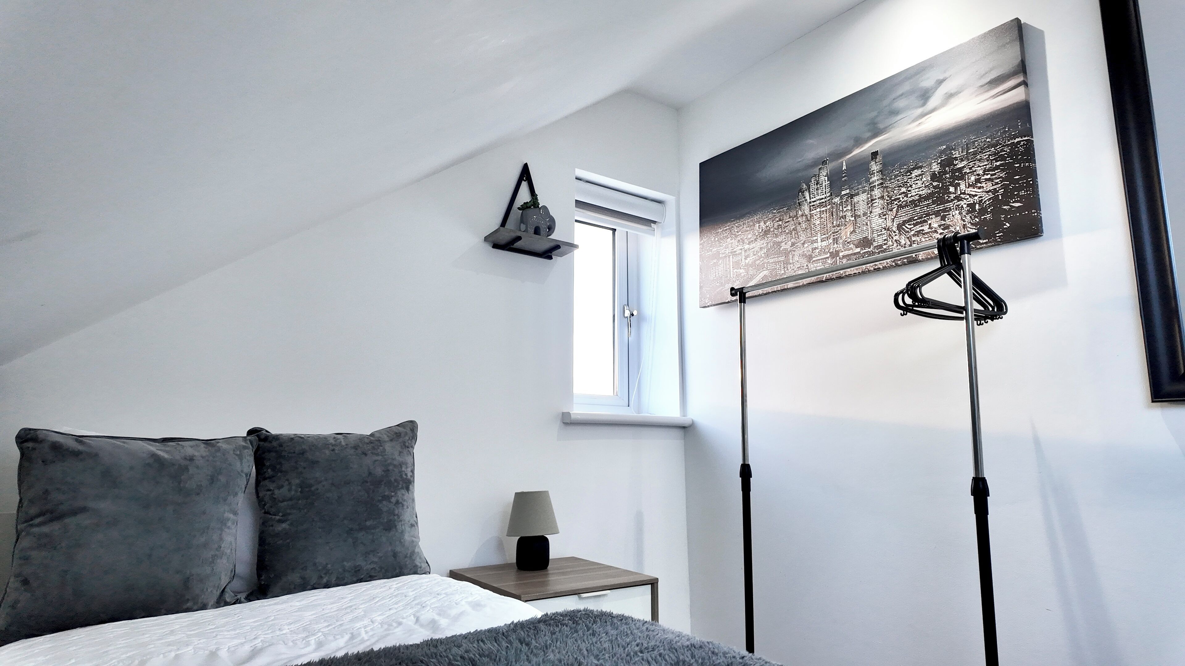 A bedroom with a bed, a nightstand, a lamp, a window, and a cityscape painting.