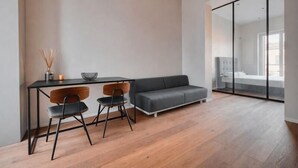 Une chambre moderne avec un plancher en bois, un canapé gris, une table à manger avec deux chaises et une cloison en verre donnant sur un espace aménagé pour dormir.