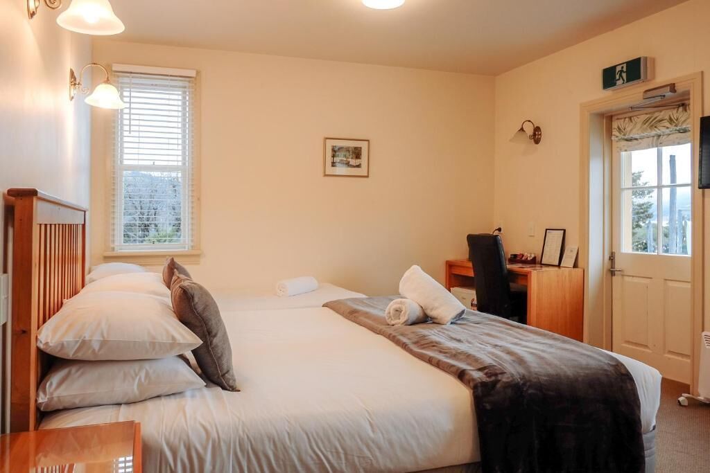 A bedroom with a bed, a desk, and a chair. There is a window with blinds, a door, and a small picture on the wall.