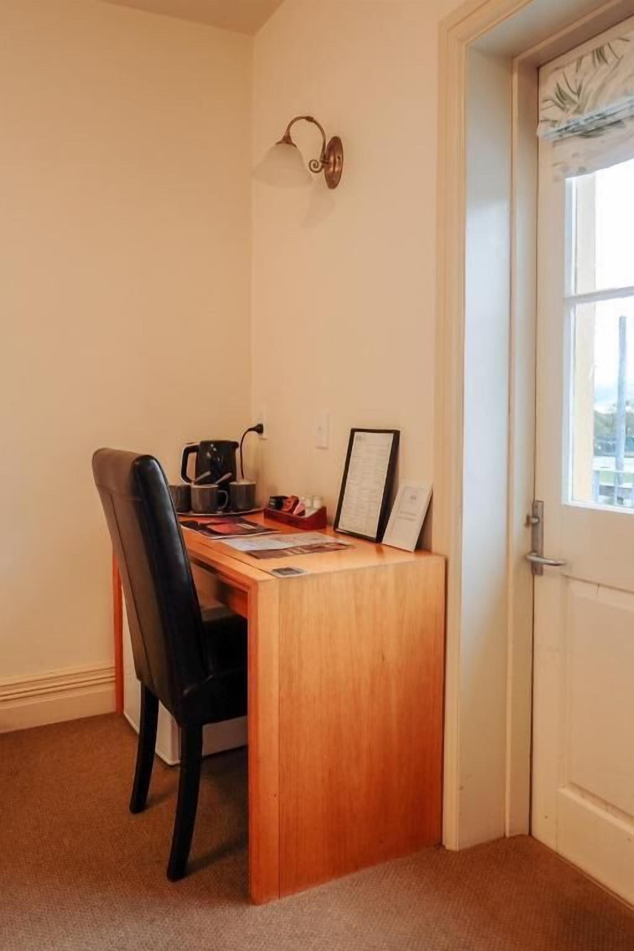 A small, neatly arranged room with a wooden desk, a black chair, a wall-mounted light fixture, and a door with a window.