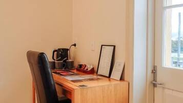 A small, neatly arranged room with a wooden desk, a black chair, a wall-mounted light fixture, and a door with a window.