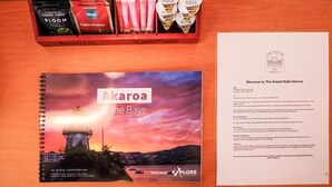A brochure for The Grand Hotel Akaroa The Bays, a spiral-bound booklet with a scenic sunset view, and a tray with instant coffee and milk packets. - The Grand Hotel & Bar (Akaroa)