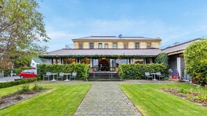 A building with a paved pathway leading to a covered entrance, surrounded by greenery and outdoor seating. - The Grand Hotel & Bar (Akaroa)