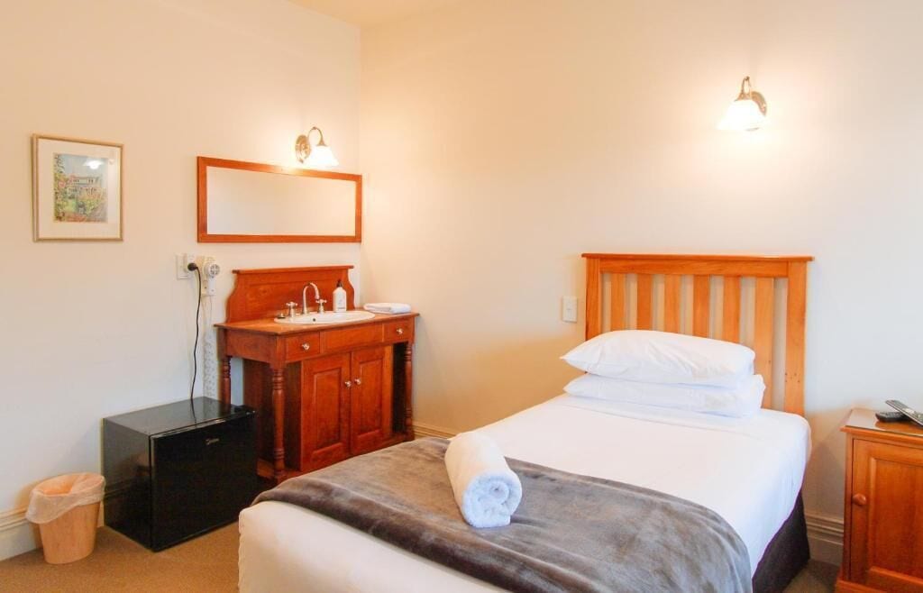 A hotel room with a bed, a wooden headboard, a nightstand, a dresser, a mirror, and a wall-mounted light fixture.