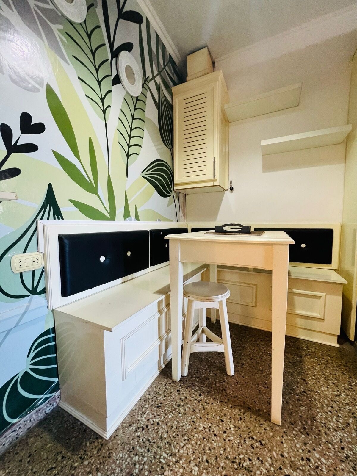 Una habitación pequeña y luminosa con un escritorio blanco, un taburete y un armario. Las paredes están decoradas con un diseño de hojas.