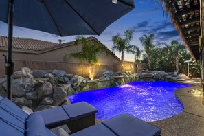 A swimming pool with a blue water feature, surrounded by rocks and palm trees, with outdoor seating and a building in the background. - Private pool & hot tub - 4mi to Polo Grounds (La Quinta)