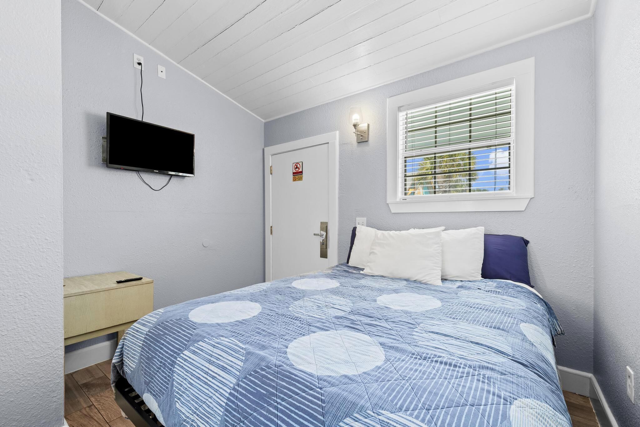 A bedroom with a bed, a television mounted on the wall, a window with blinds, and a door with a sign.