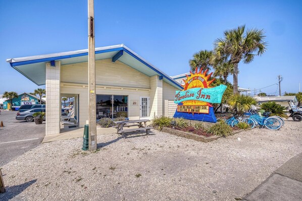 Un motel frente a la playa con un letrero que dice 'Paradise Inn', un acceso cubierto y un área de estacionamiento con algunos vehículos.