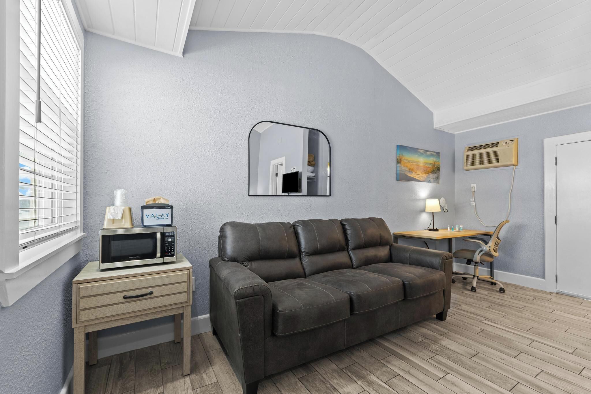 Une pièce comprenant un canapé en cuir foncé, une petite table de chevet en bois avec un micro-ondes, un miroir, un bureau avec une lampe et une chaise.