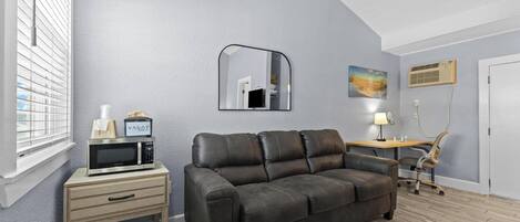 Une pièce comprenant un canapé en cuir foncé, une petite table de chevet en bois avec un micro-ondes, un miroir, un bureau avec une lampe et une chaise.