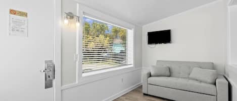 A room with a white door, a window with blinds, a grey sofa, and a mounted TV.
