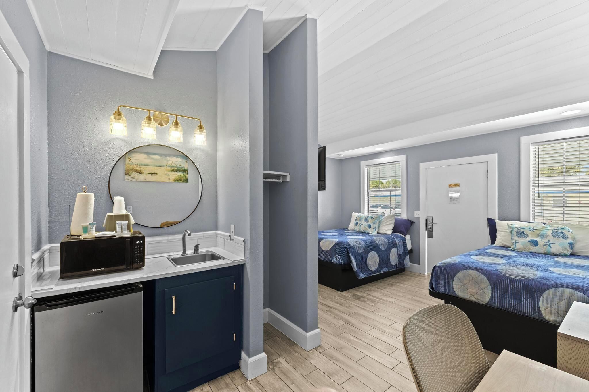 Een compacte hotelkamer met kitchenette, een bed met een blauw gebedecoreerd laken, een klein zithoekje en een groot raam met zonwering.