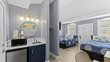 A compact hotel room with a kitchenette, a bed with a blue patterned bedspread, a small seating area, and a large window with blinds.