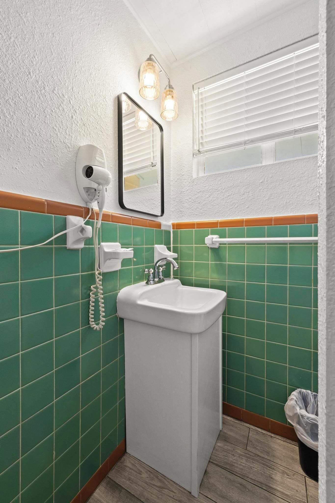 Une salle de bain avec un lavabo blanc, des murs recouverts de carreaux verts, un sèche-cheveux et un miroir.
