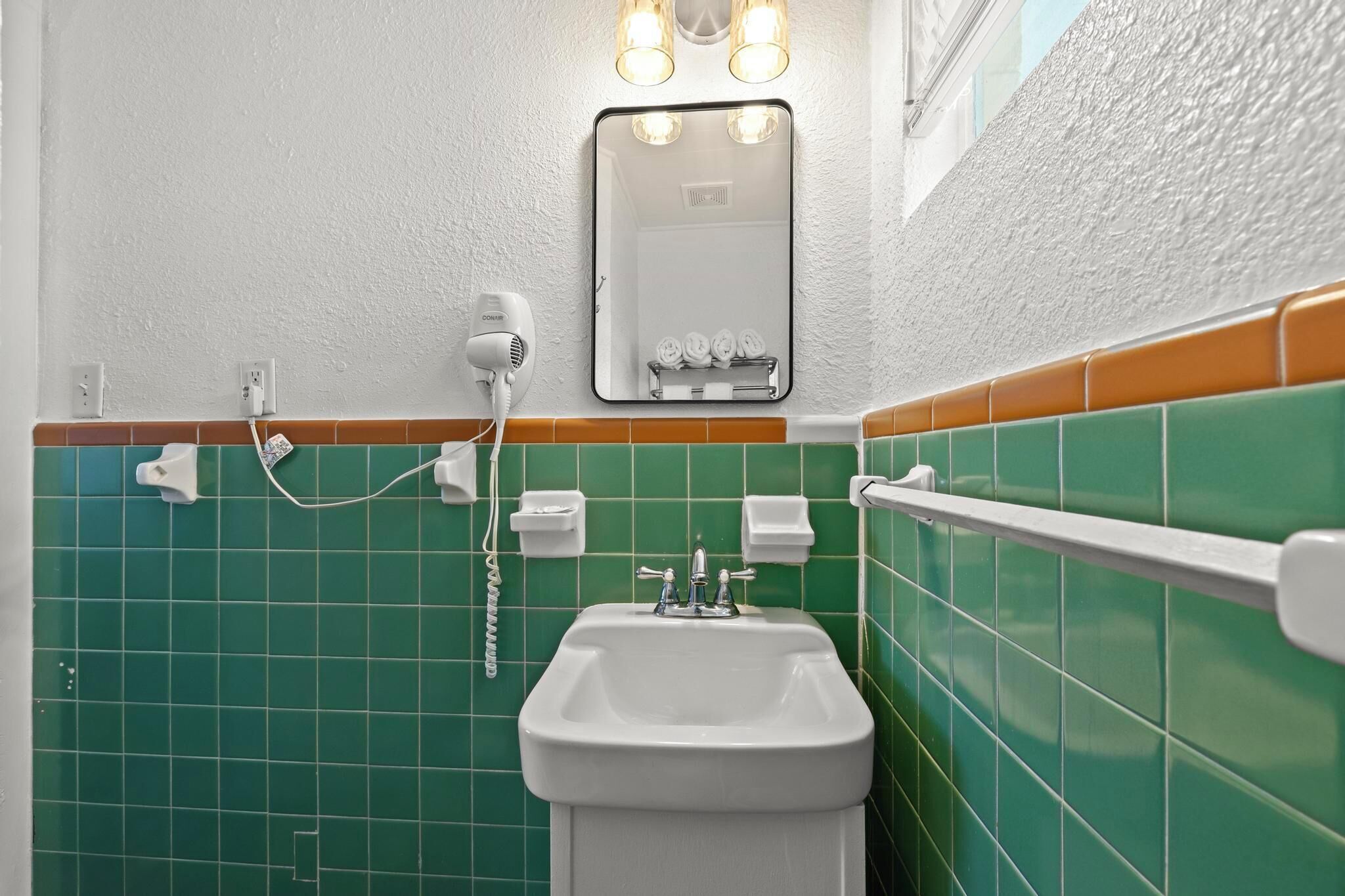 A bathroom with a white sink, green tiles, a mirror with lights, and a hairdryer.