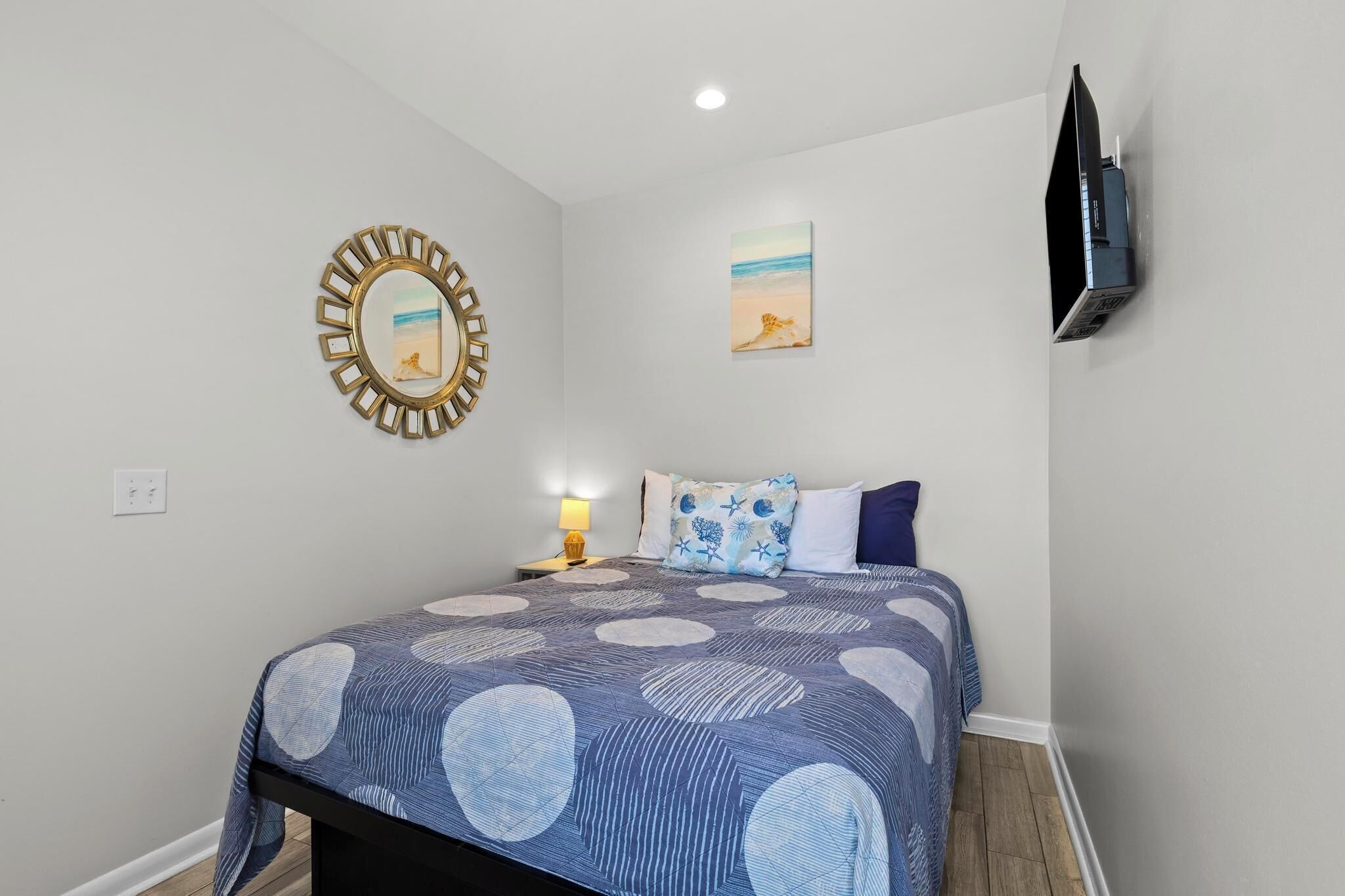 A bedroom with a bed, a wall-mounted TV, a framed picture, and a bedside lamp.