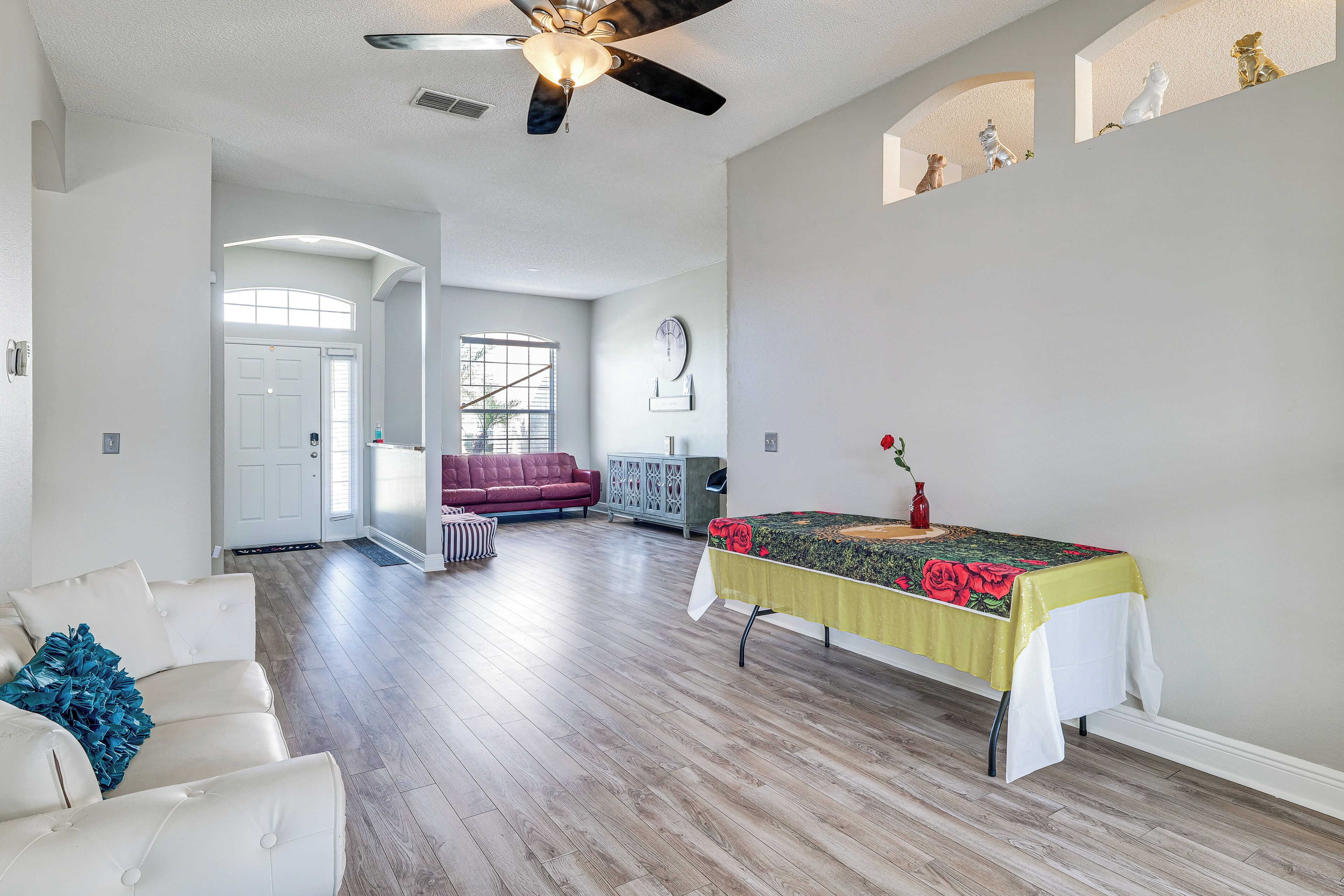 Amplia sala de estar con ventilador de techo alto, un sofá rojo, una mesa con mantel floral y vista a una puerta que da a otra habitación.