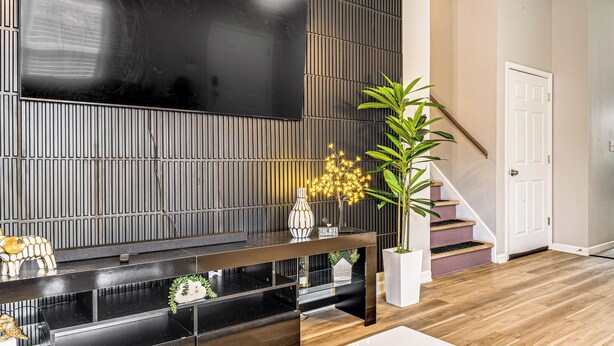 Ein modernes Wohnzimmer mit einem großen Flachbildfernseher an einer dunklen Wand, einem Konsolentisch mit Dekoration, einer Treppe und einer Topfpflanze.
