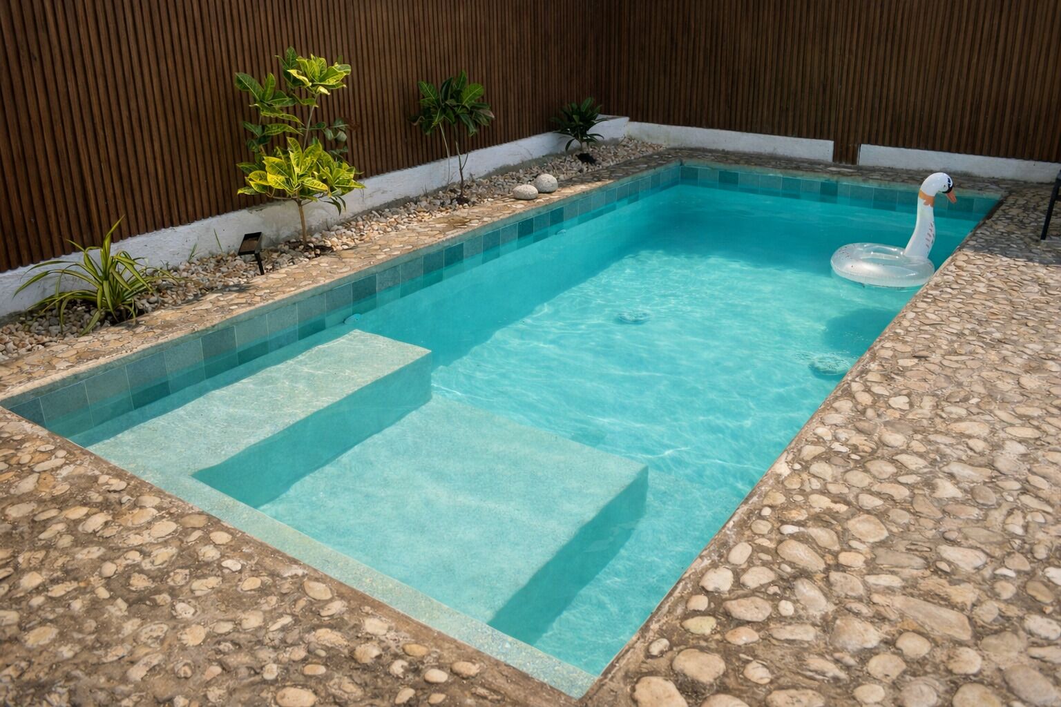 A rectangular swimming pool with clear blue water, surrounded by a stone deck and a wooden fence.