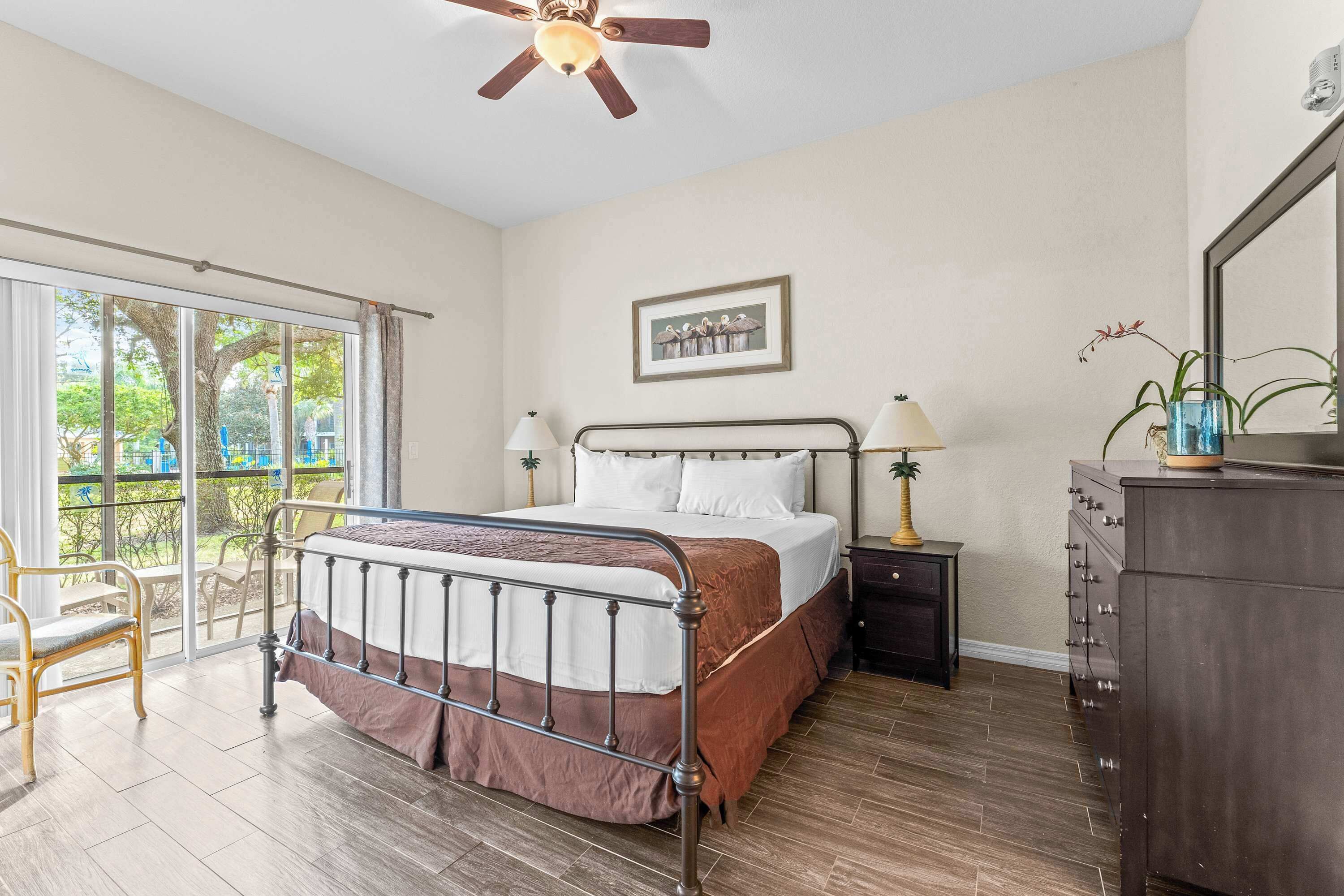 Une chambre à coucher comprenant un lit, une table de chevet, une commode, un ventilateur de plafond, une fenêtre avec vue et un tableau encadré au mur.