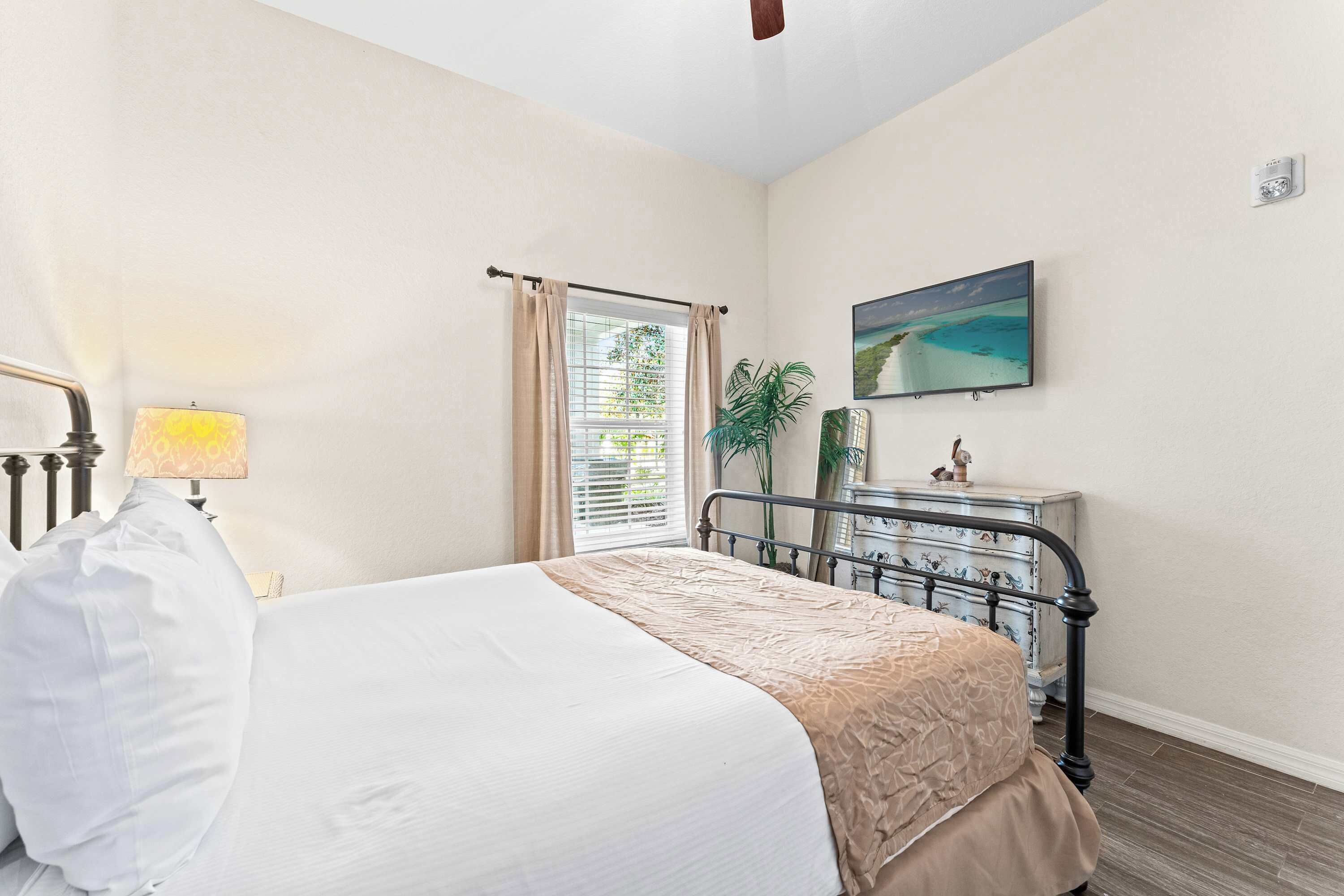 A bedroom with a bed, a bedside lamp, a window with curtains, a dresser, a mirror, and a painting on the wall.