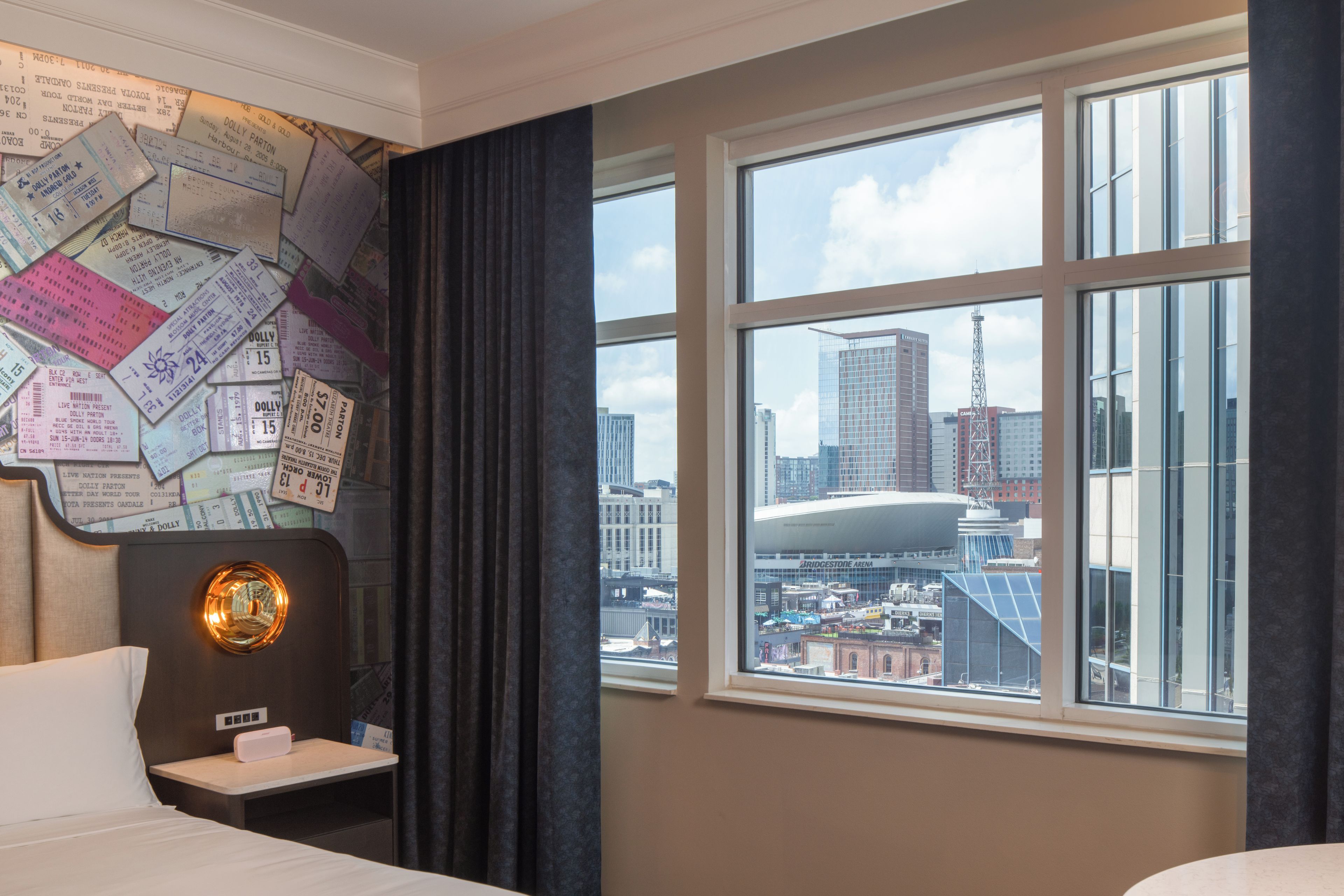 A hotel room with a large window offering a view of a cityscape, a wall covered with various papers, and a bedside table with a lamp.