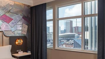 A hotel room with a large window offering a view of a cityscape, a wall covered with various papers, and a bedside table with a lamp.
