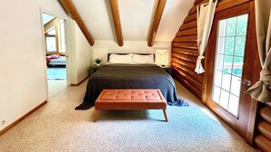 Ein Schlafzimmer mit schrägen Wänden, Holzbalken, einem Bett mit dunkler Decke, einem Nachttisch mit Lampe und einem Fenster mit Vorhängen.