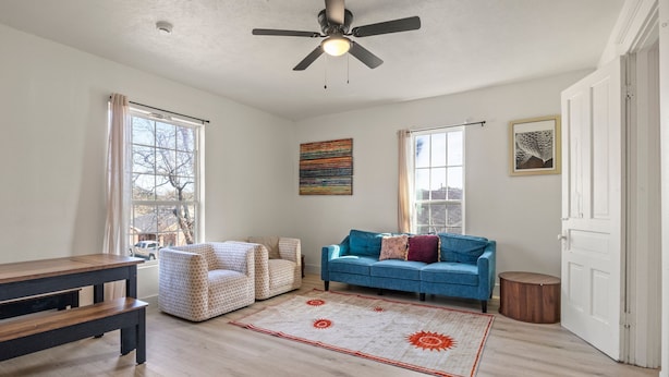 Ein Wohnzimmer mit einem blauen Sofa, gemusterten Sesseln, einem Holztisch und einem weißen Teppich mit Sonnenmotiven in Rot. Eine Deckenventilator ist ebenfalls zu sehen.