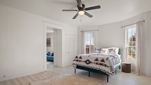 Ein Schlafzimmer mit einem Bett, einem Deckenventilator, einem Fenster mit Vorhängen und einer Tür zu einem anderen Raum.