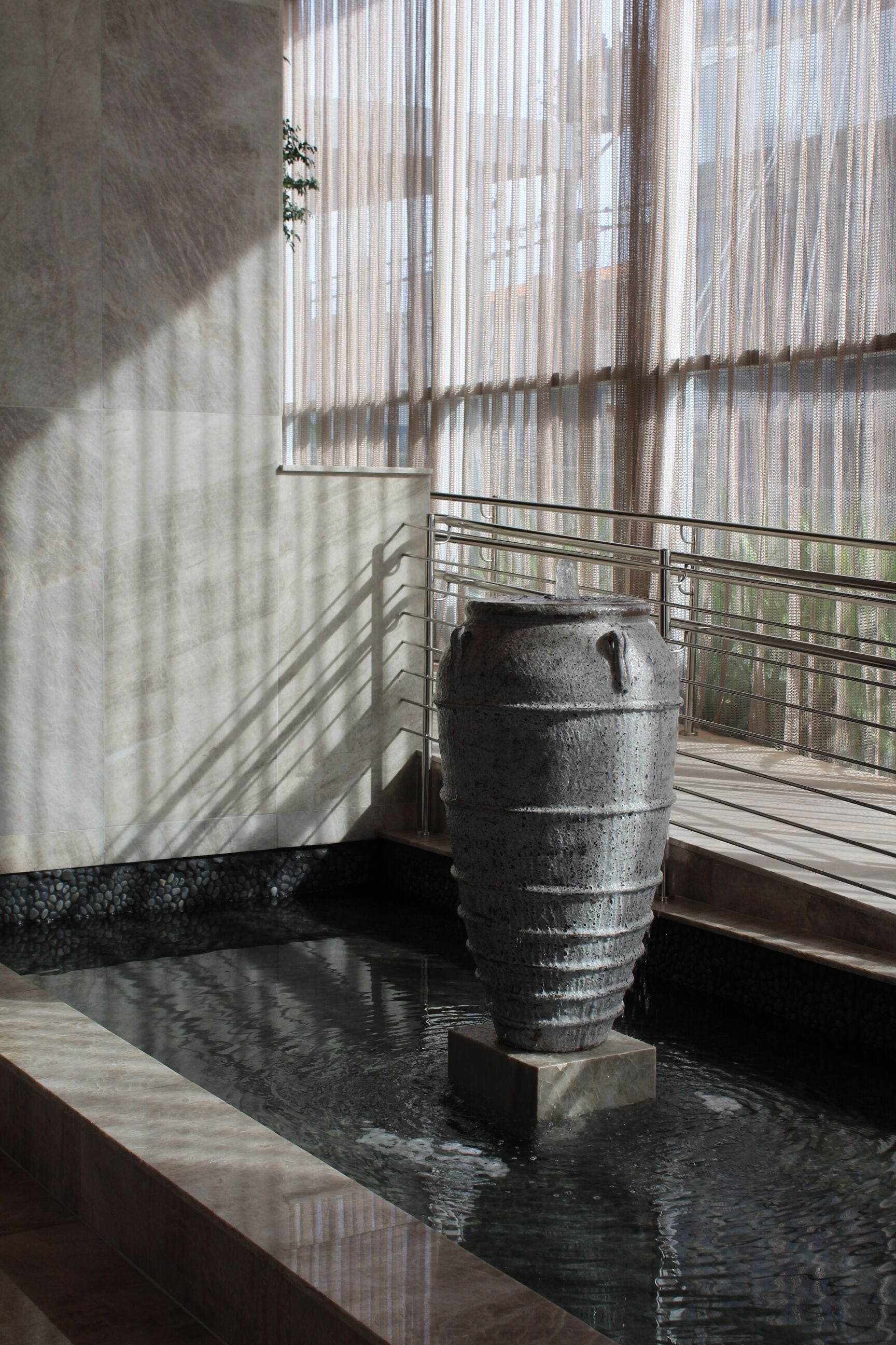 A stone water feature in a modern indoor setting with a metal railing and a view of trees through sheer curtains.