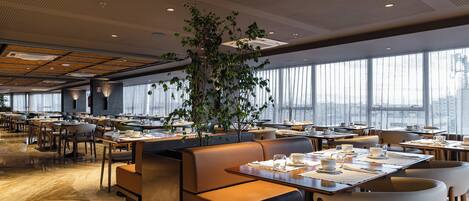 A modern restaurant with high ceilings, large windows, and a central plant. Tables are set with white tableware.
