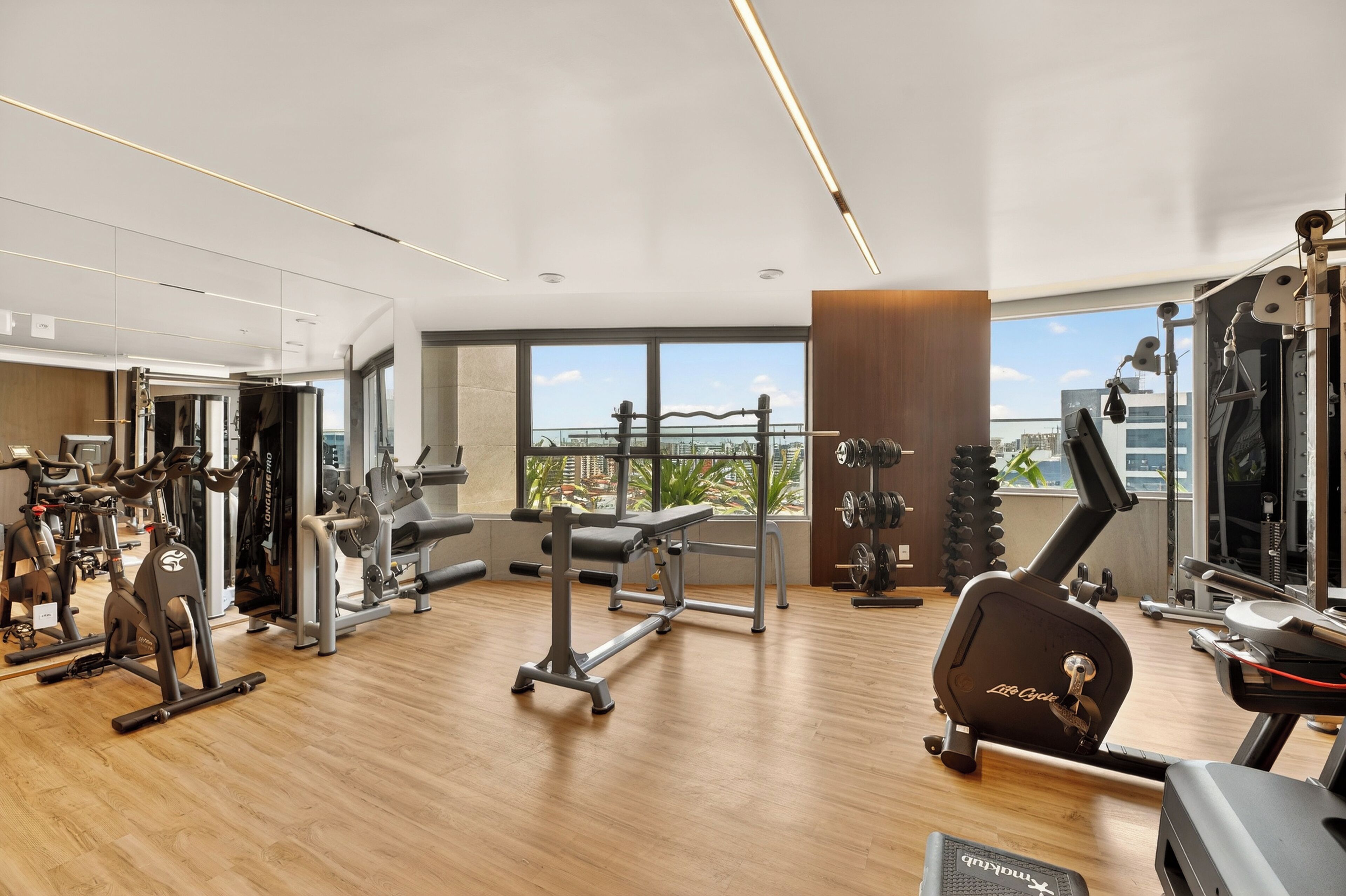 A modern gym with various exercise equipment, large windows, and a view of the city.