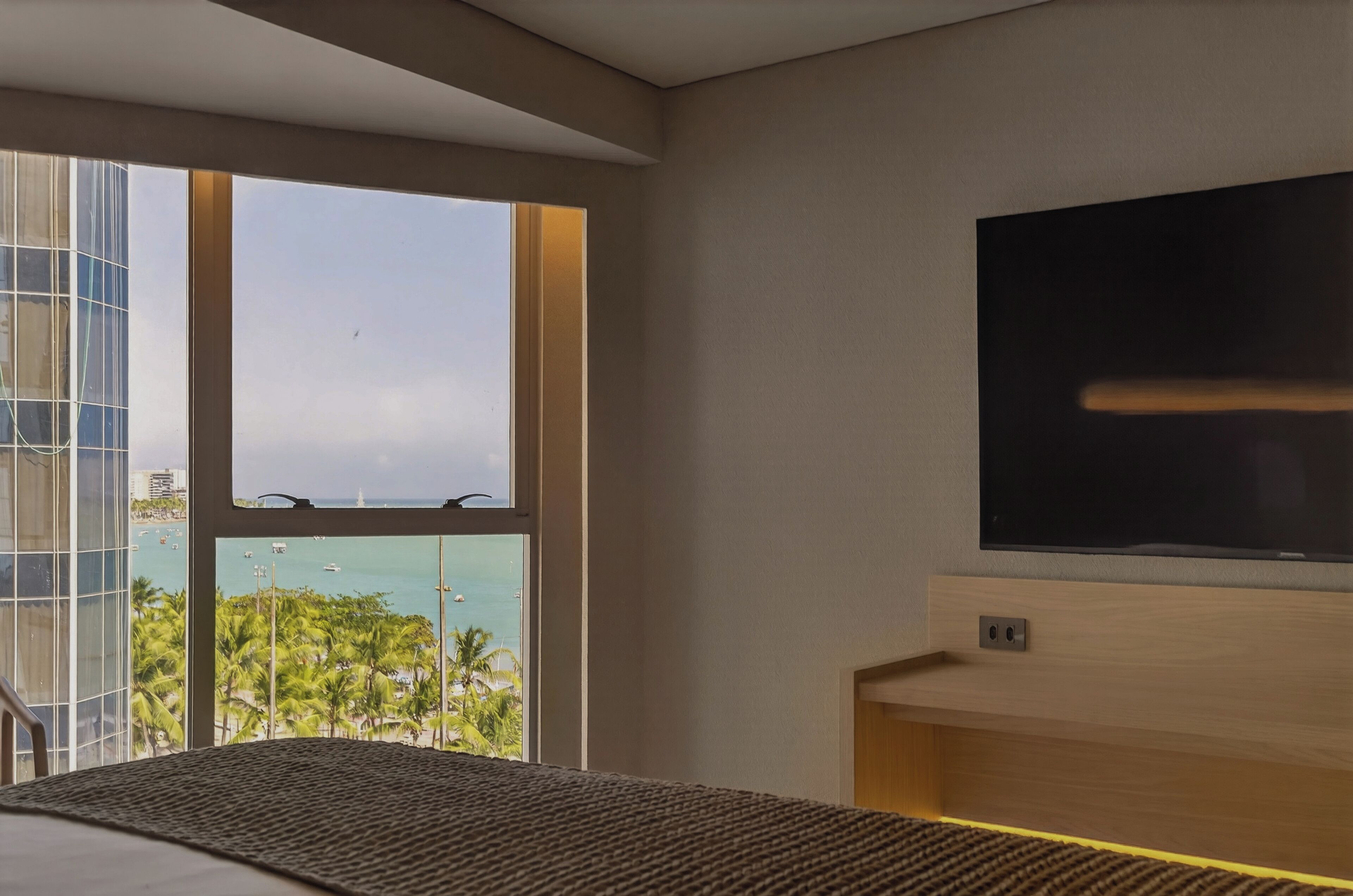 Habitación de hotel con un ventanal que ofrece vistas al océano y palmeras, un televisor de pantalla plana montado en la pared y una cama con cabecera de fibras naturales.