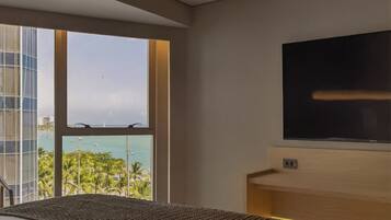 Habitación de hotel con un ventanal que ofrece vistas al océano y palmeras, un televisor de pantalla plana montado en la pared y una cama con cabecera de fibras naturales.