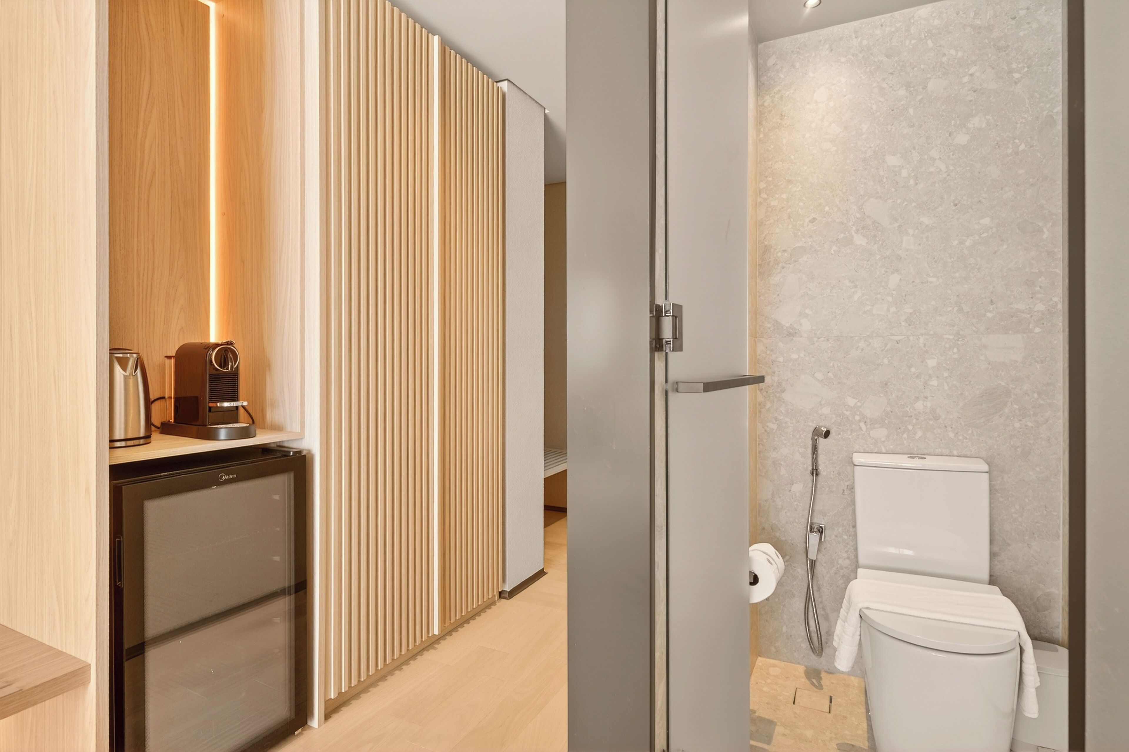 A compact kitchenette with a coffee maker and a mini-fridge, a sliding wooden partition, and a modern bathroom with a marble wall and a toilet.
