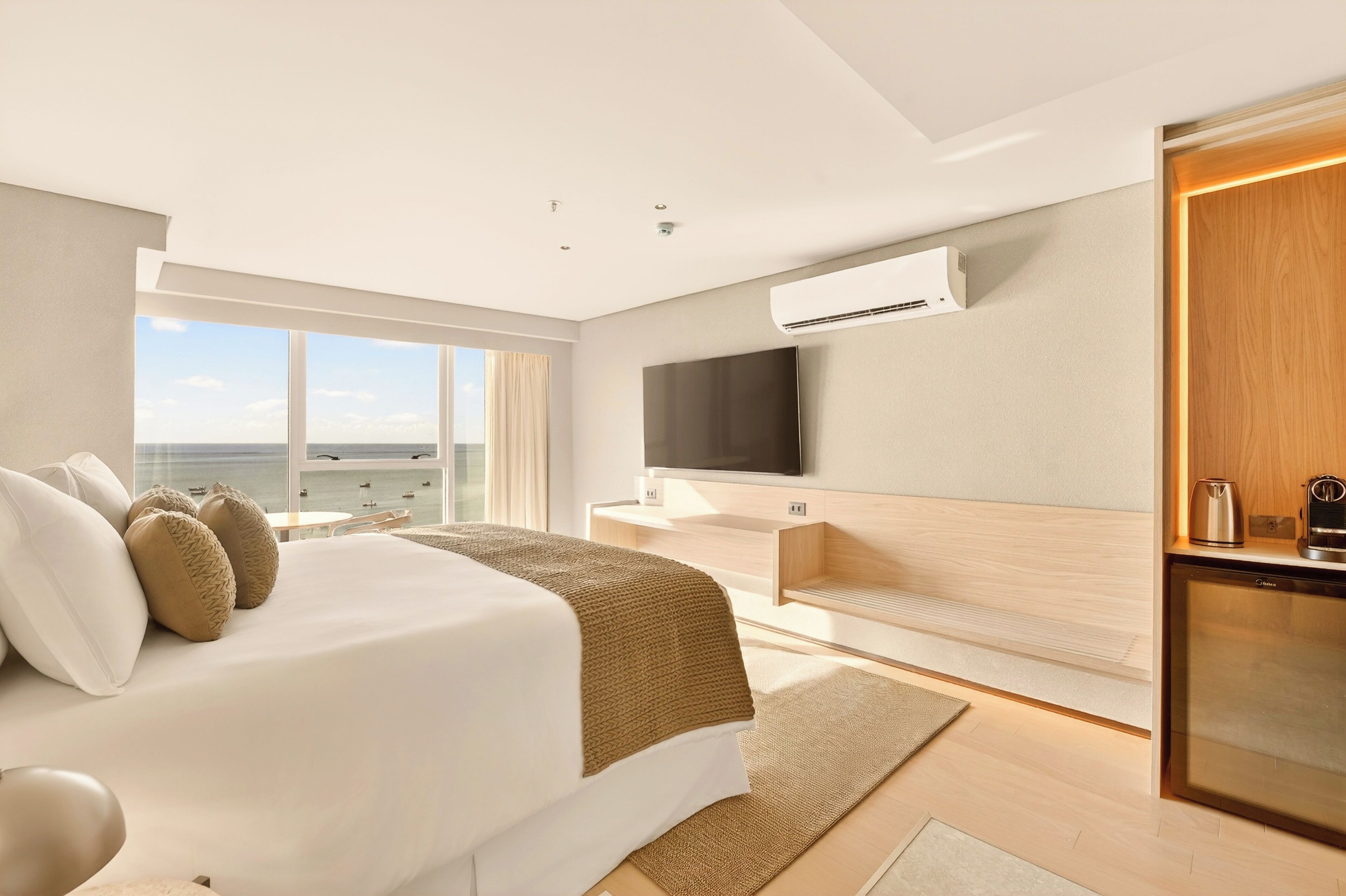 A modern hotel room with a large bed, a flat-screen TV, an air conditioning unit, and a view of the sea.