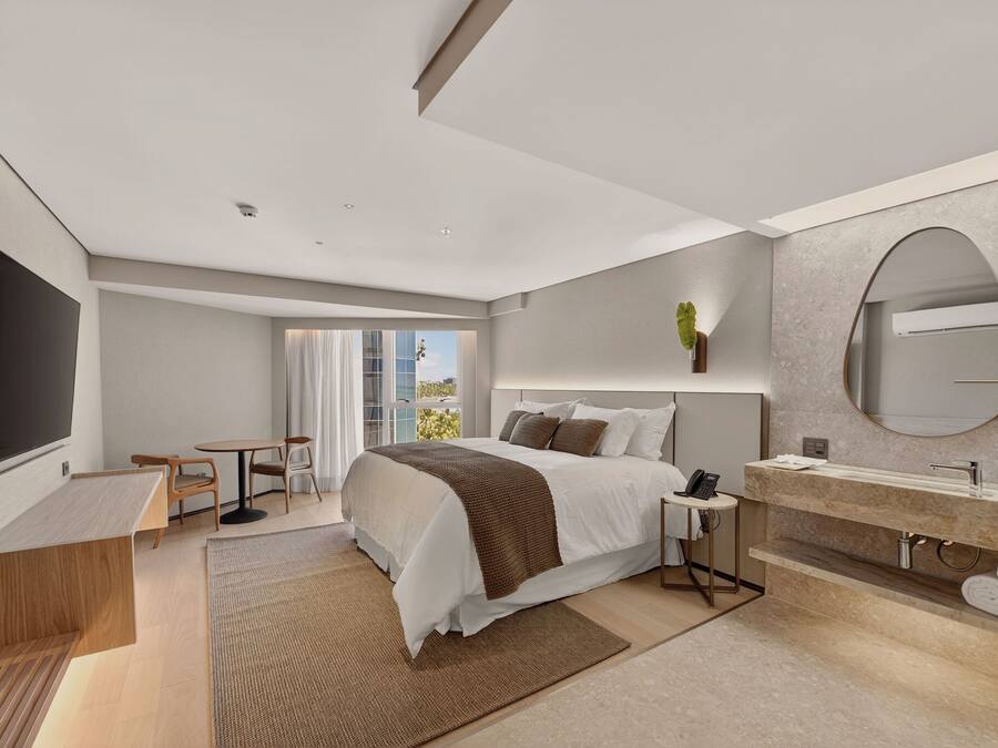A modern hotel room with a large bed, a TV, a desk, a bathroom with a mirror, and a view of the outdoors.