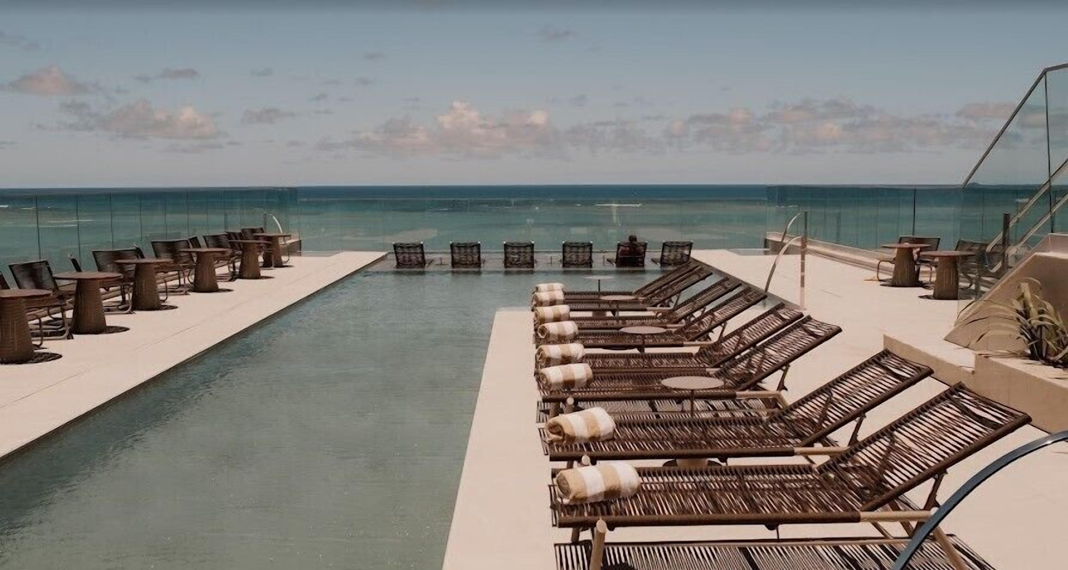 Un área junto a la piscina con sillones y mesas, vista despejada al océano y una barrera de vidrio.